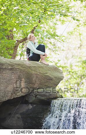 Stock Image - USA, New York, New York City, Manhattan, Central Park, Young woman sitting on rock. Fotosearch