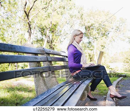 Stock Photo - USA, New York, New York City, Manhattan, Central Park, Young woman sitting on bench and using laptop. Fotosearch
