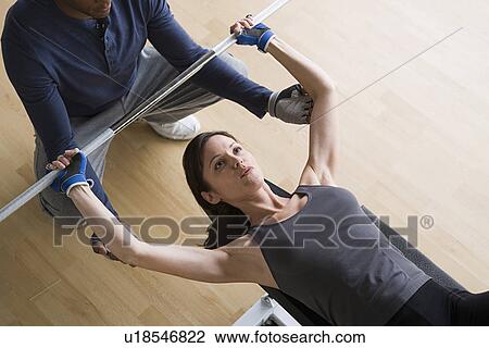 Woman lifting barbell while instructor assisting her View Large Photo Image Stock Image - Woman lifting barbell while instructor assisting her. Fotosearch