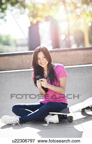 Stock Photo - Young chinese woman text messaging while sitting on skateboard. Fotosearch