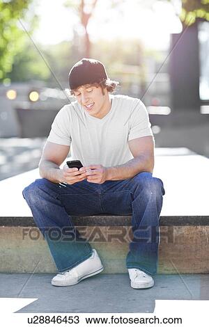 Young man sitting outdoors, text messaging View Large Photo Image Stock Image - Young man sitting outdoors, text messaging. Fotosearch