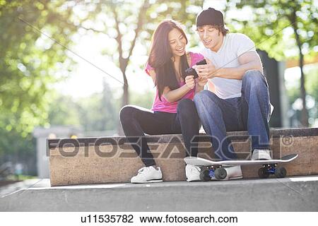 Stock Image - Young multi-racial couple sitting with skateboard, using mobile phone. Fotosearch