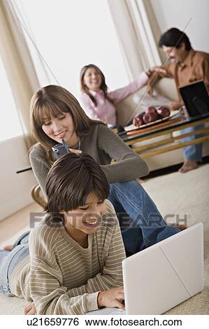 Stock Photograph - Brother and sister using laptop and mobile, parents in background. Fotosearch