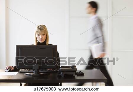 Stock Image - Businesswoman working on computer, businessman walking in background. Fotosearch