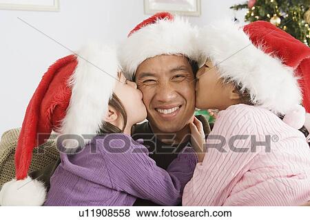 Stock Photo - daughters (10-11) kissing father wearing santa hat. Fotosearch