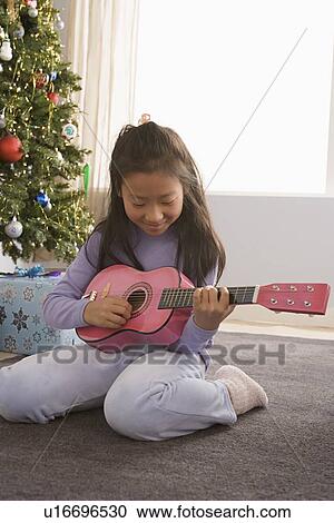 Stock Image - Girl (10-11) playing guitar, Christmas tree in background. Fotosearch