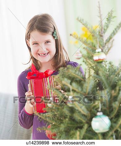 Stock Photo - Girl (8-9) standing with gift behind Christmas tree. Fotosearch