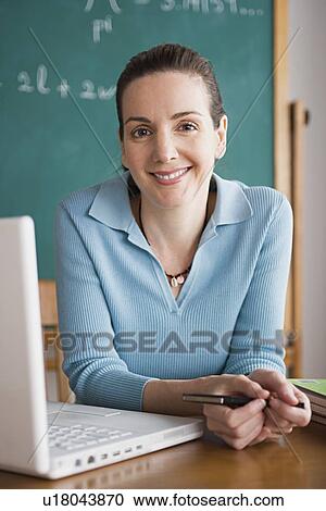 Stock Image - Happy female teacher posing with laptop while sitting in classroom. Fotosearch