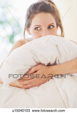 Stock Image - Portrait of young woman embracing pillow in bedroom. Fotosearch