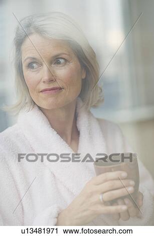 Stock Image - Senior woman looking through window in morning. Fotosearch
