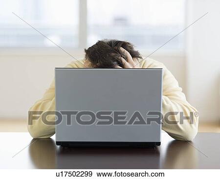Stock Photo - Stressed man sitting at desk behind laptop with head in hands. Fotosearch