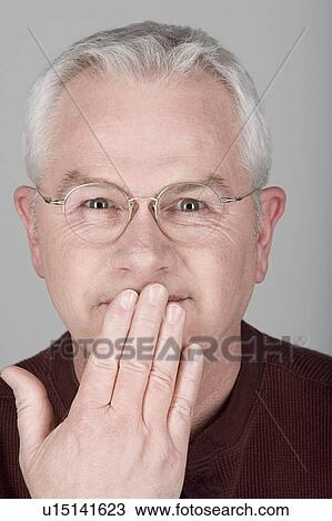 Stock Image - Studio shot portrait of senior man with hand covering mouth, close-up. Fotosearch