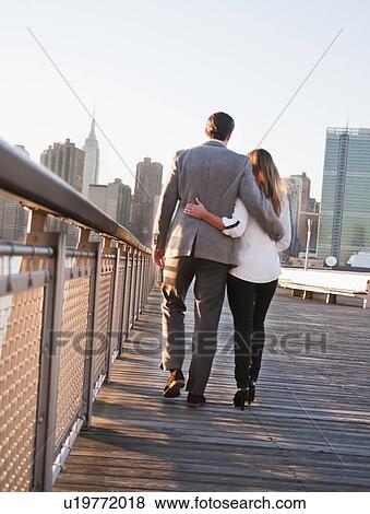 Stock Photo - USA, New York, Long Island City, Rear view of young couple walking on boardwalk, Manhattan skyline in background. Fotosearch