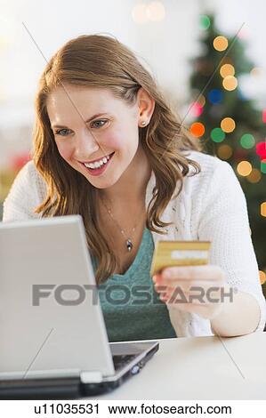 Stock Image - Young woman on-line shopping from home with Christmas tree on background. Fotosearch