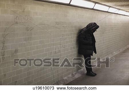 Stock Image - Hooded person leaning on subway wall. Fotosearch