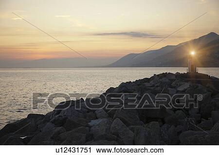 Stock Image - Lighthouse shining over rocky beach. Fotosearch