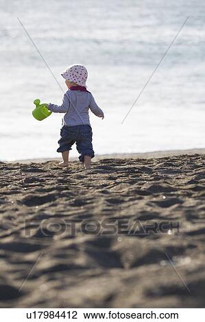 Stock Image - Toddler carrying watering can on beach. Fotosearch