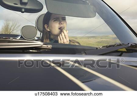Stock Image - Woman applying lipstick in car mirror. Fotosearch