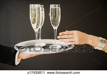 Stock Image - Close up of man's and woman's hands and tray with champagne flutes, studio shot. Fotosearch