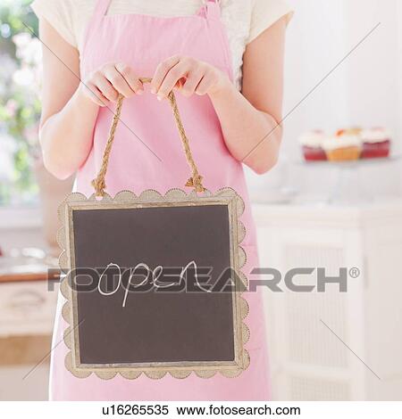 Stock Photography - Close up of young woman in apron holding open sign. Fotosearch