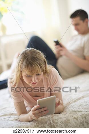 Couple lying on bed and using digital devices View Large Photo Image Stock Photo - Couple lying on bed and using digital devices. Fotosearch