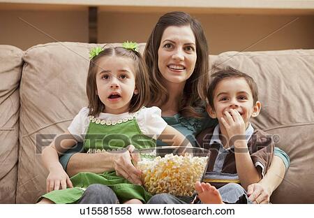 Stock Photo - Front view of mother with children (4-5, 6-7) sitting on couch watching tv and eating popcorn. Fotosearch