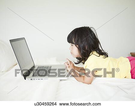 Picture - Girl lying down in bed, using laptop (4-5). Fotosearch