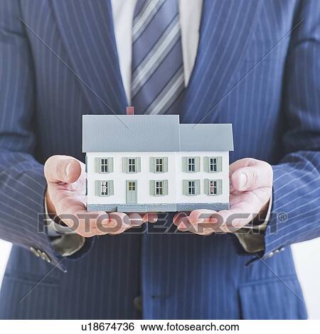 Stock Photograph - Man wearing suit holding small house model. Fotosearch