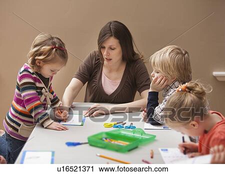 Stock Image - Mid adult woman teaching children (2-3, 4-5) drawing in kindergarten. Fotosearch