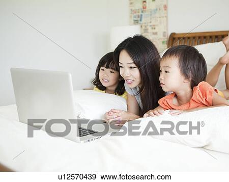 Stock Photo - Mother with daughters (2-3, 4-5) lying down in bed, using laptop. Fotosearch