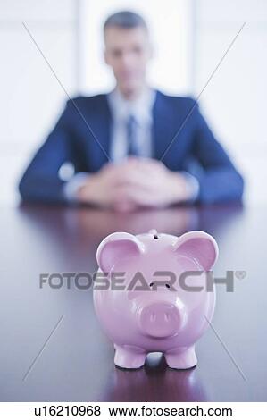 Piggybank on desk, businessman sitting on background View Large Photo Image Stock Photo - Piggybank on desk, businessman sitting on background. Fotosearch