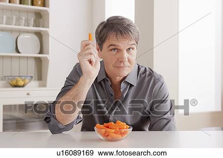 Stock Photo - Portrait of mature man with baby carrot on table. Fotosearch