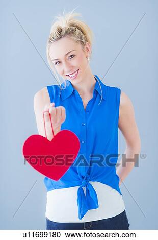 Stock Image - Portrait of smiling young woman holding red heart, studio shot. Fotosearch