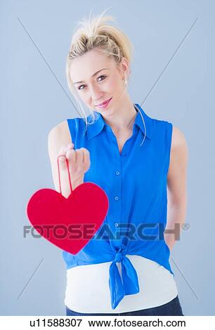 Stock Photo - Portrait of smiling young woman holding red heart, studio shot. Fotosearch