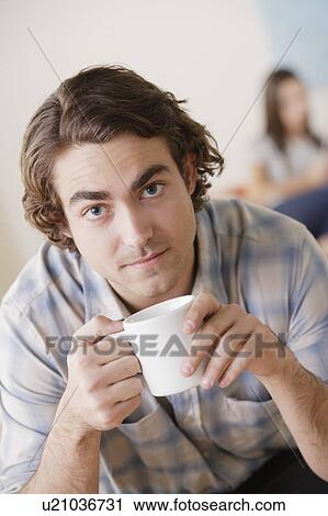 Stock Image - Portrait of young man holding mug, woman in background. Fotosearch