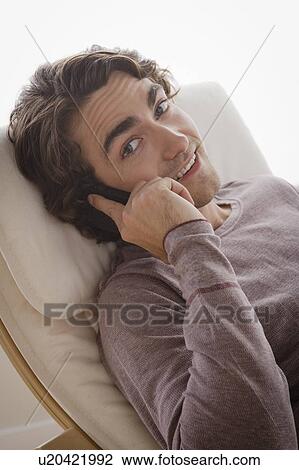 Stock Image - Portrait of young man sitting on armchair and talking via mobile. Fotosearch