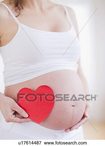 Stock Photo - Pregnant woman holding red paper heart on her belly. Fotosearch