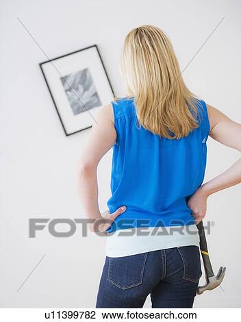Stock Image - Rear view of woman holding hammer and looking at painting on wall. Fotosearch