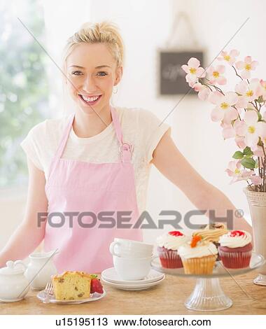 Stock Image - Smiling young woman in apron standing at table with cakes. Fotosearch