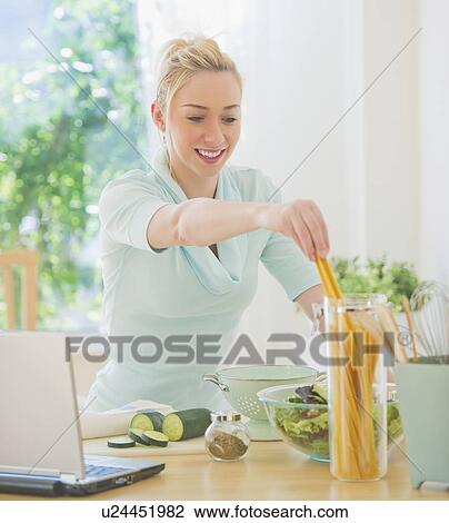 Stock Image - Smiling young woman preparing food in kitchen. Fotosearch