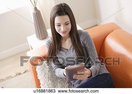 Stock Image - Smiling young woman using digital tablet on sofa. Fotosearch