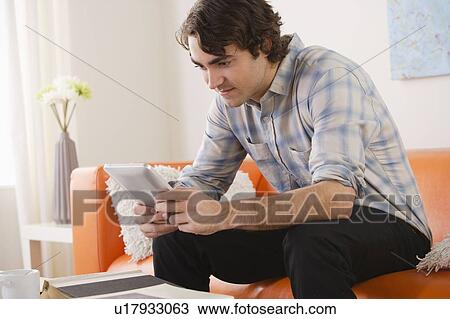Stock Image - Young man sitting on sofa and using digital tablet. Fotosearch