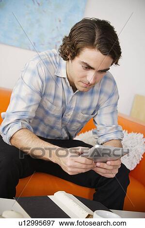 Young man sitting on sofa and using digital tablet View Large Photo Image Stock Photo - Young man sitting on sofa and using digital tablet. Fotosearch