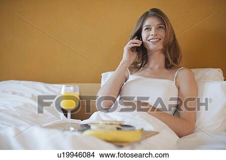 Picture - Young woman calling friend while enjoying breakfast in bed. Fotosearch