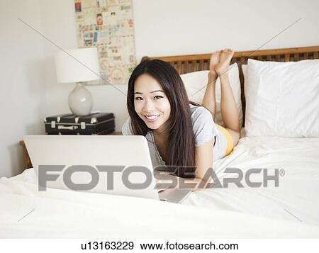 Stock Photo - Young woman lying down in bed, using laptop. Fotosearch