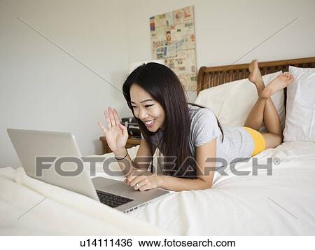 Young woman lying down in bed, using laptop, waving View Large Photo Image Stock Photograph - Young woman lying down in bed, using laptop, waving. Fotosearch