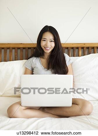 Young woman sitting on bed, using laptop View Large Photo Image Stock Image - Young woman sitting on bed, using laptop. Fotosearch
