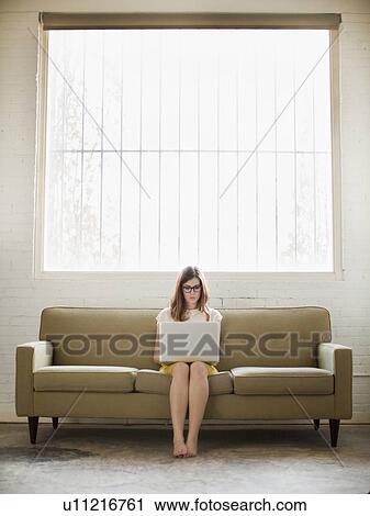 Stock Image - Young woman sitting on sofa with laptop. Fotosearch