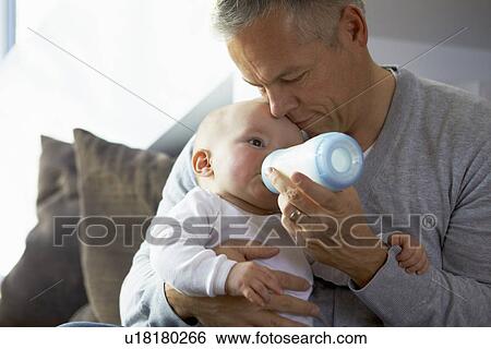 Stock Photograph - Father bottle feeding baby on sofa. Fotosearch