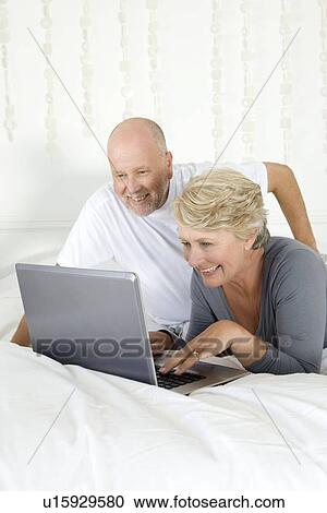 Stock Image - Older couple using laptop on bed. Fotosearch
